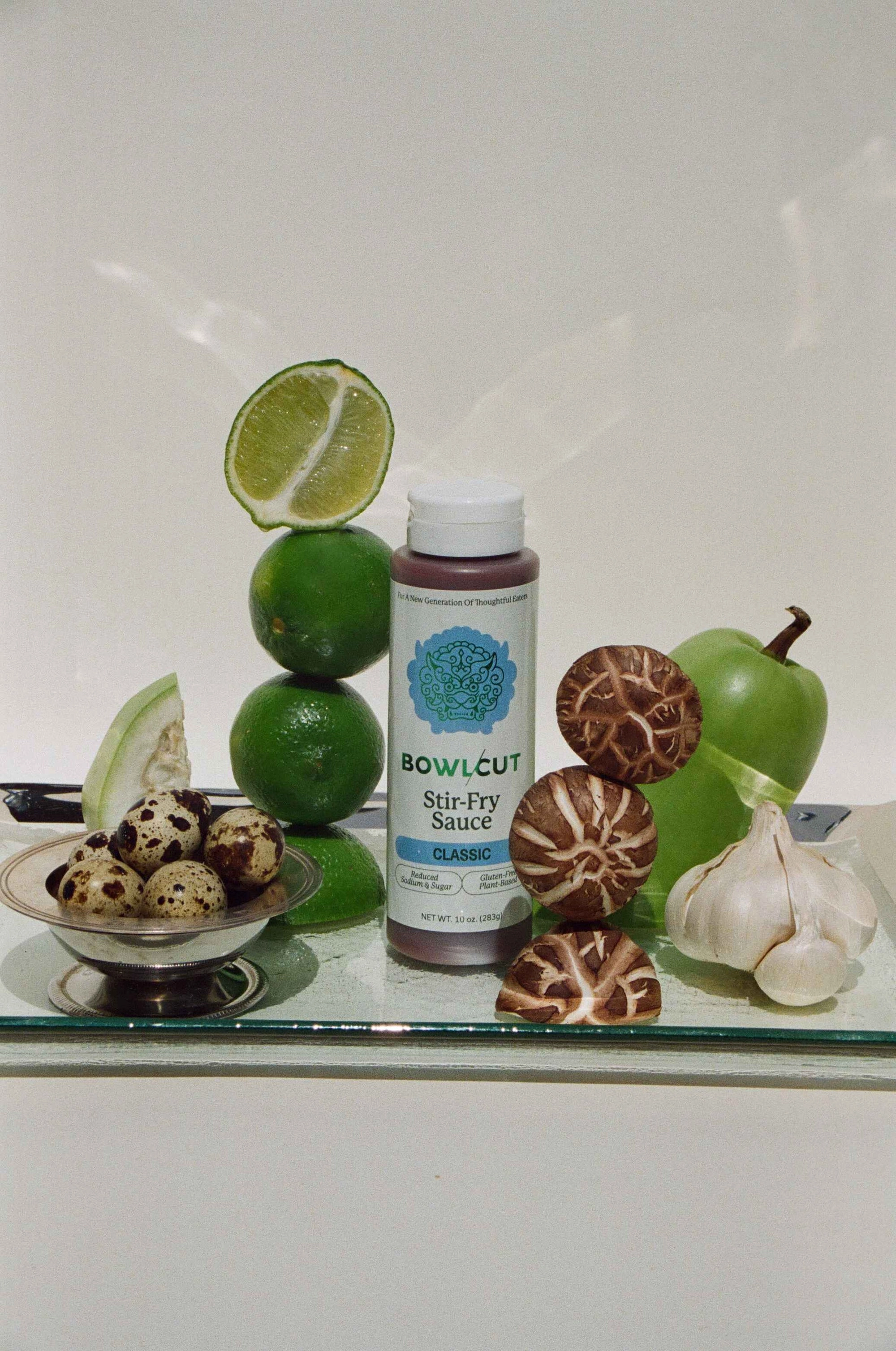 Bottle of 'Bowlcut Stir-Fry Sauce' surrounded by limes, mushrooms, and garlic on a reflective surface.