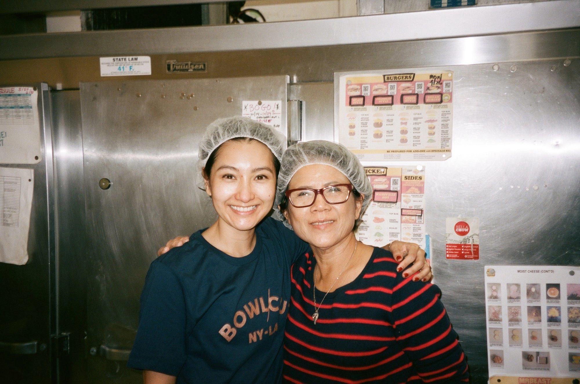 Two people in hairnets and navy Bowlcut shirts smiling in a stainless-steel kitchen